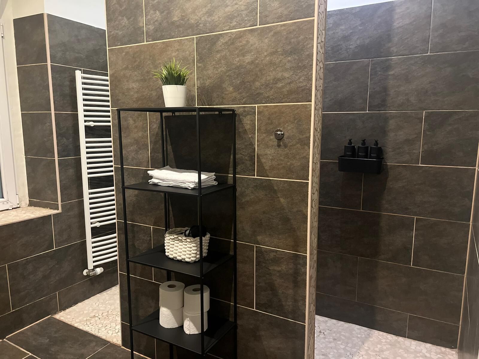 Un baño con paredes de azulejos oscuros, un estante de metal negro con toallas, una planta y artículos de aseo.