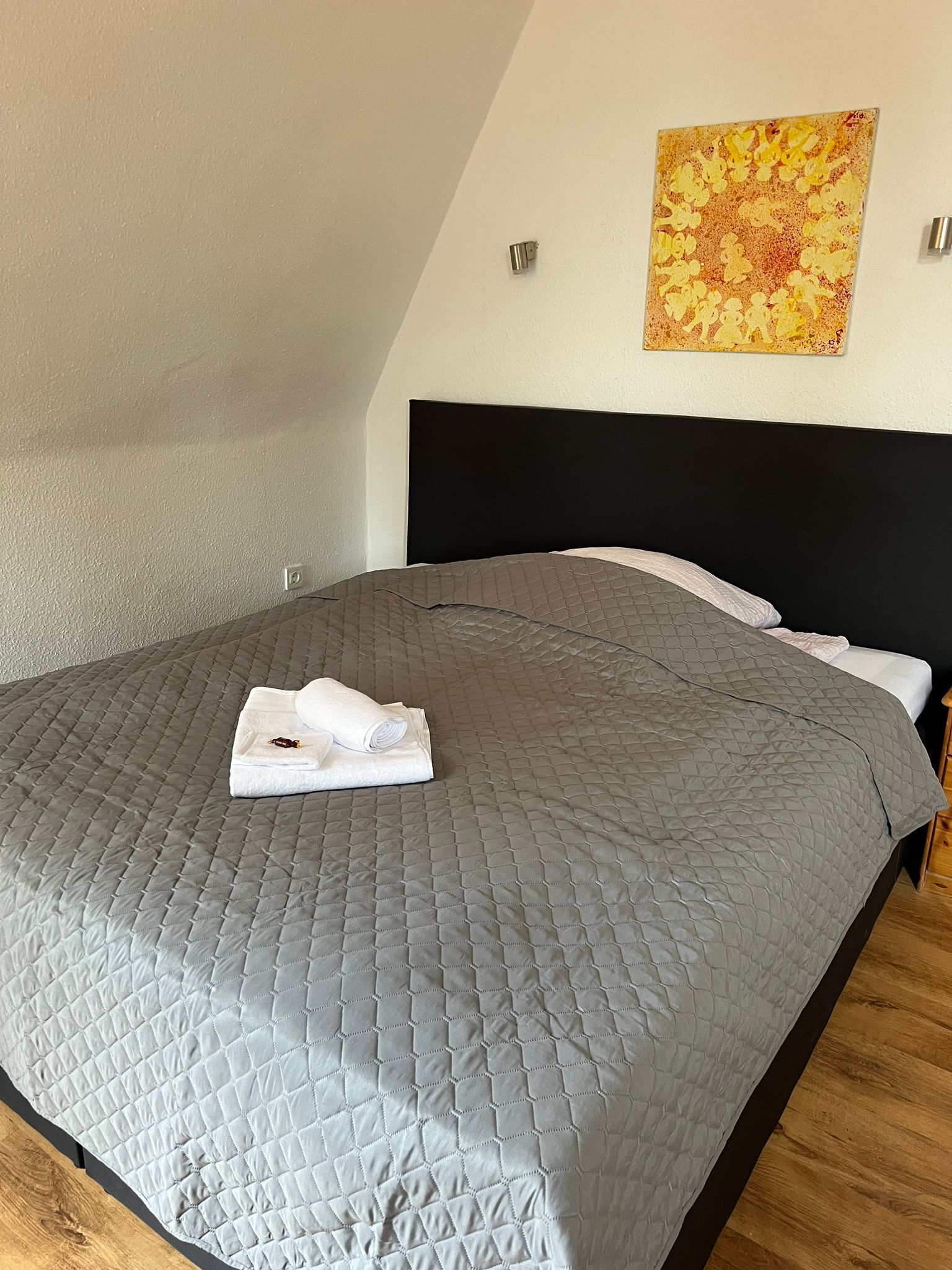 Een net opgemaakt bed met een grijze quilt, een gevouwen witte handdoek en een schilderij aan de muur.