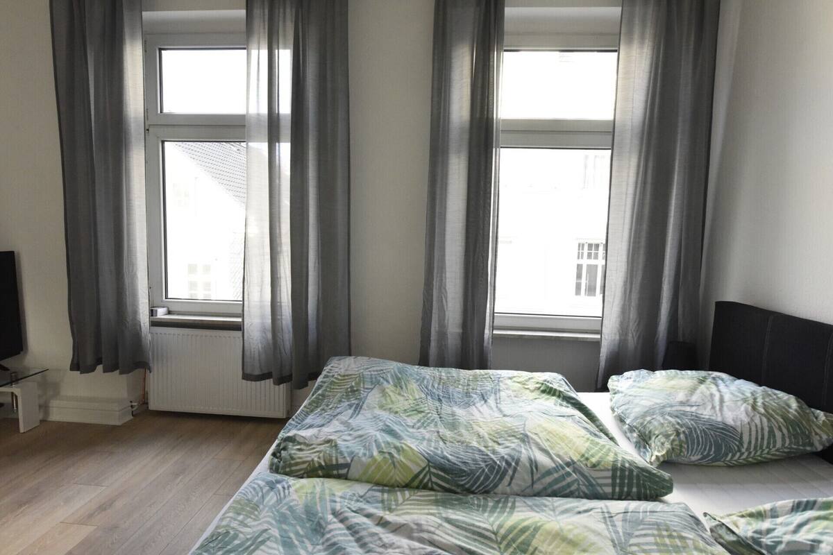 A bedroom with a bed, two windows with curtains, a radiator, and a television.