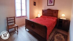 Une chambre à coucher avec un lit en bois, une chaise, une fenêtre avec des rideaux, une table de chevet avec une lampe et un tableau encadré au mur.