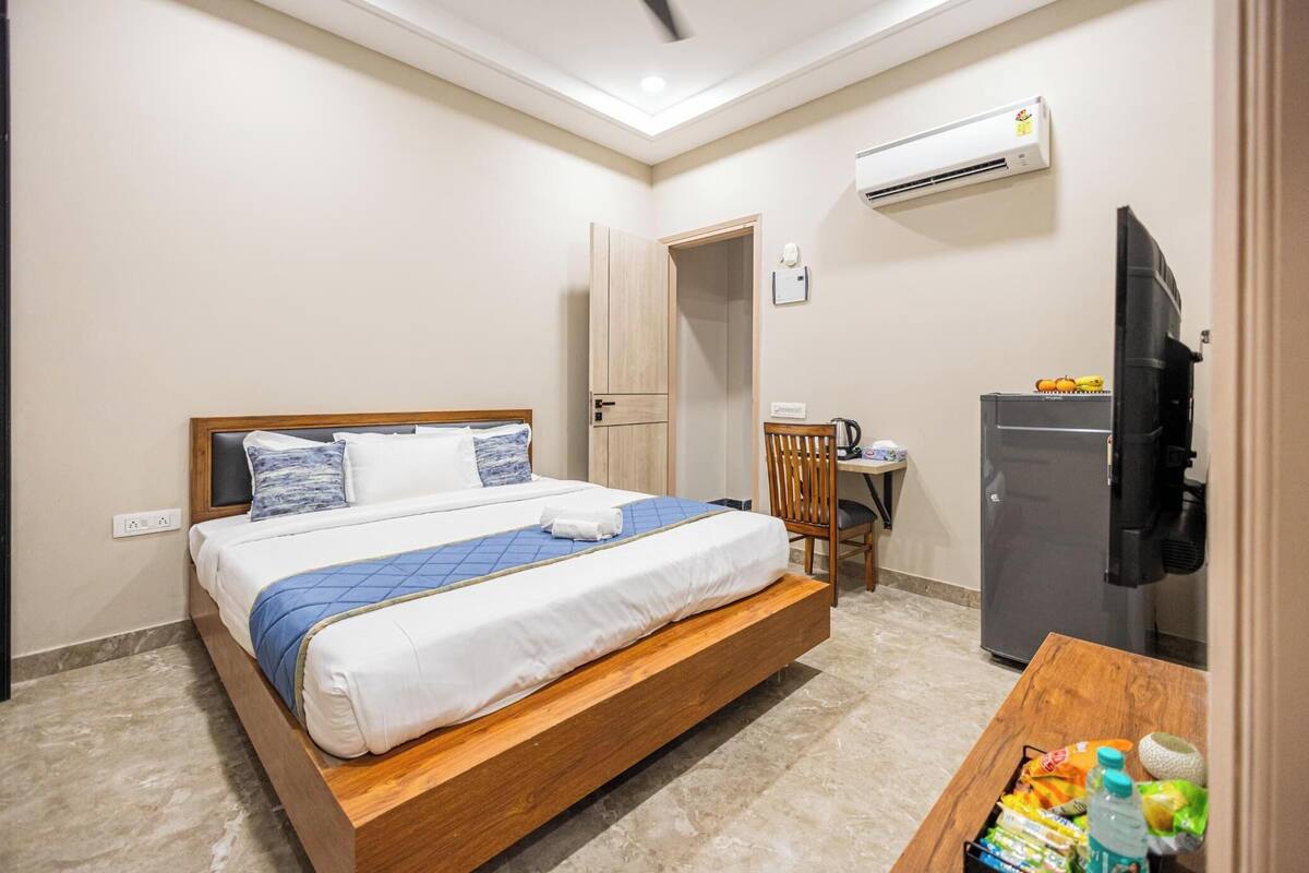 A hotel room with a bed, a desk with a chair, a refrigerator, and an air conditioning unit.