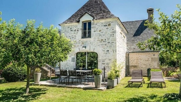 A stone building with a pitched roof, a small porch with a table and chairs, and a grassy yard with two lounge chairs. - Gite de la Bouysse Haute (CUZANCE)