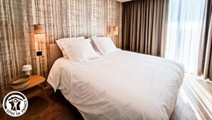 Een hotelkamer met een groot bed, twee bedlampjes, een houten nachtkastje en een raam met gordijnen.