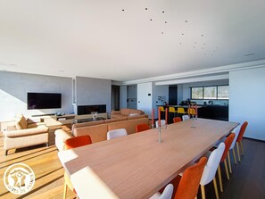 Una sala de estar moderna con una larga mesa de comedor de madera, sillas anaranjadas y blancas, un televisor de pantalla plana y una zona de cocina con barra.