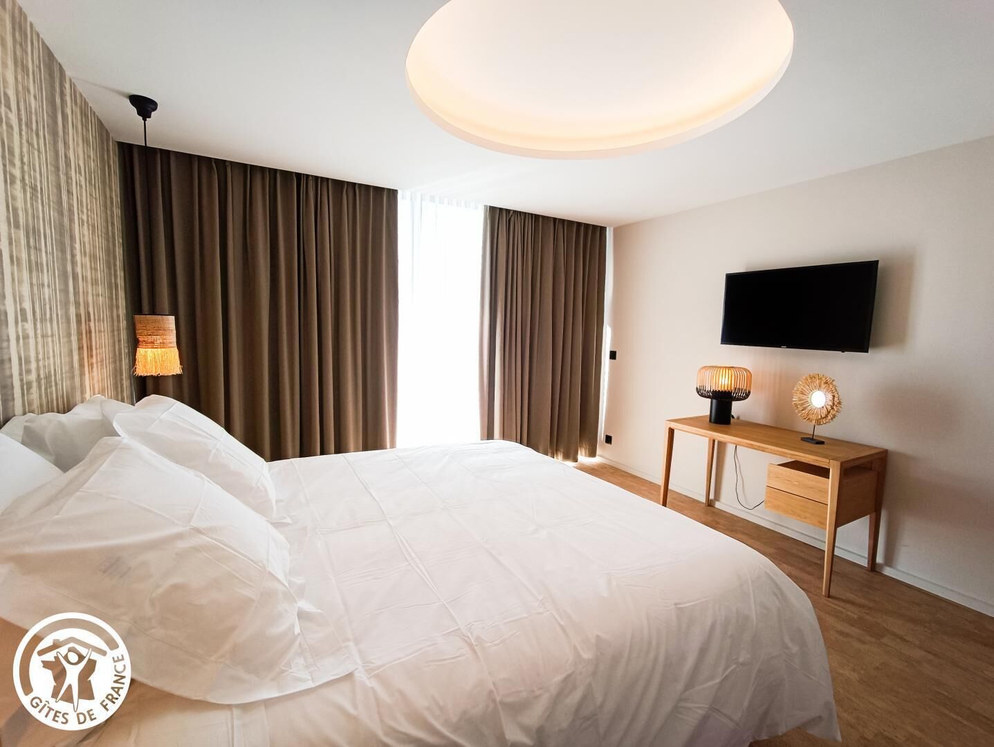 Une chambre d’hôtel avec un grand lit, une table de chevet en bois avec deux lampes, un téléviseur à écran plat fixé au mur et une grande fenêtre avec des rideaux.