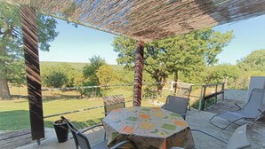 An outdoor patio area with a round table and chairs, a pergola overhead, and a view of a grassy field and trees. - Gîtes de France® - La Quercynoise (BERGANTY)