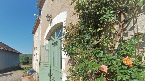A rustic building with a green door, potted plants, and blooming roses. - Gîtes de France® - La Quercynoise (BERGANTY)