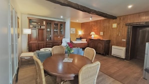 A cozy dining area with a round wooden table surrounded by wicker chairs, a small side table, and a vase with flowers. A kitchen with wooden cabinets and a radiator is visible in the background. - Gîtes de France® - La Quercynoise (BERGANTY)
