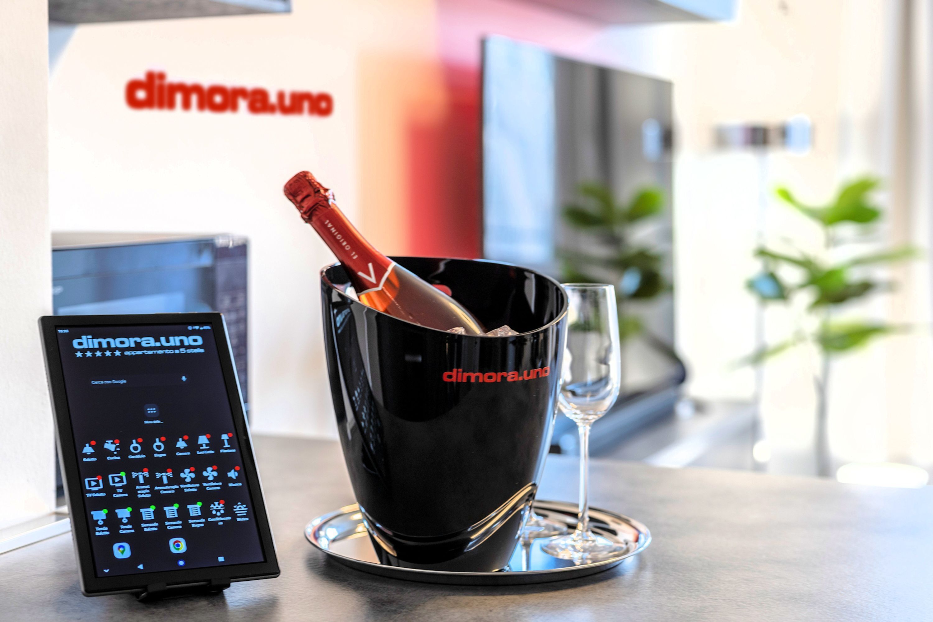 Une bouteille de champagne dans un seau noir, avec un verre à côté, un appareil numérique dont l’écran affiche « dimora.uno », et une enseigne portant le même nom en arrière-plan.
