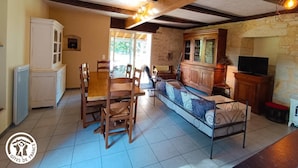 Una sala acogedora con una mesa de madera, sillas, un sofá y una televisión. Hay una zona de cocina con armarios y una pared de piedra.