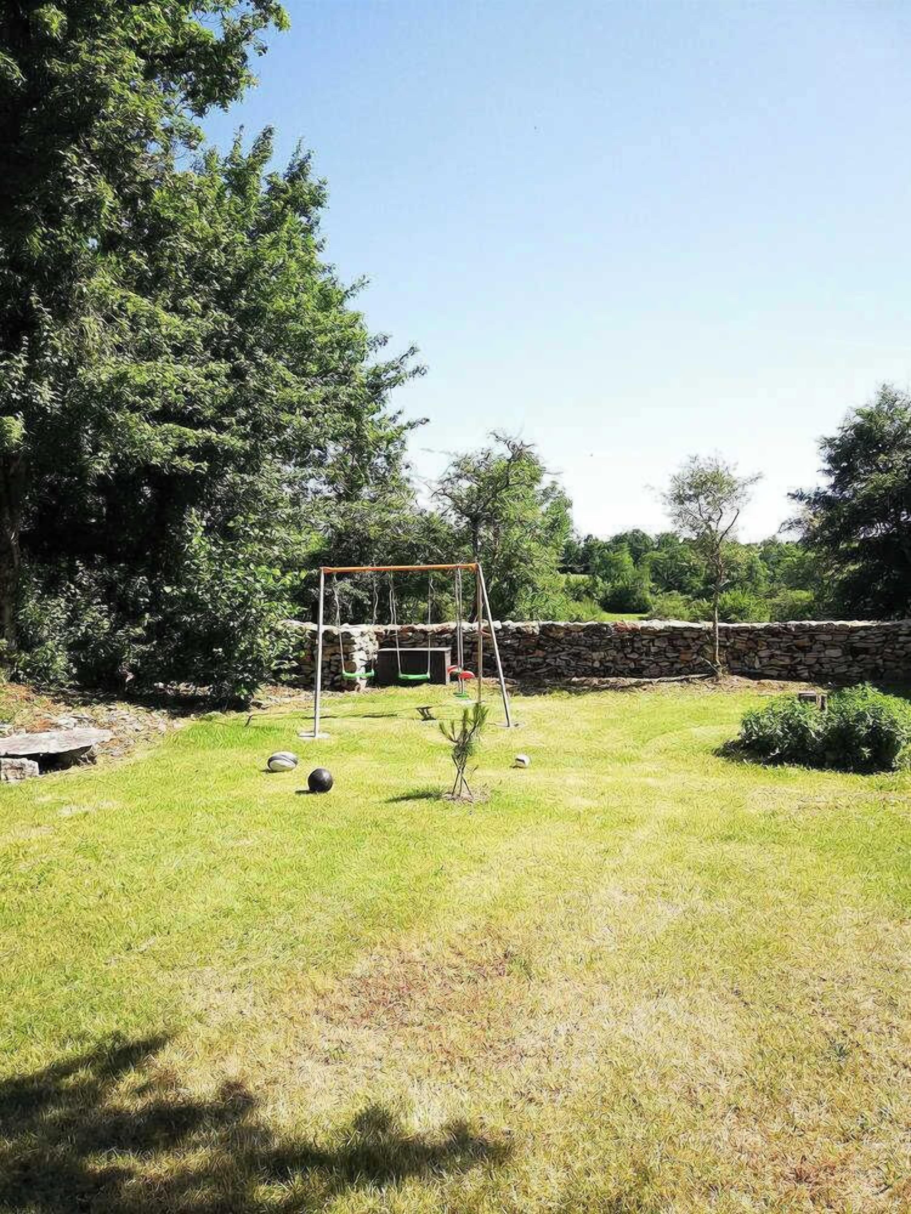 A grassy backyard with a swing set, a stone wall, and trees.