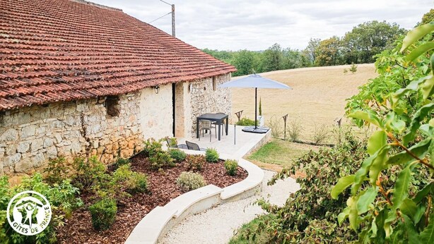 Un bâtiment en pierre avec un toit en tuiles, une terrasse avec des sièges extérieurs et une vue sur un champ.