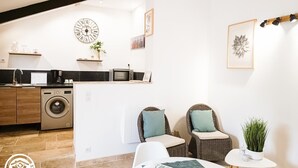 Un espace de vie compact comprenant un coin cuisine, un lave-linge, deux fauteuils en osier, une petite table et une horloge murale.