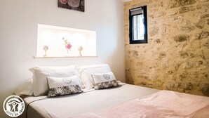 Une chambre avec un mur en pierre, un lit avec une literie blanche, une fenêtre et un tableau représentant des fleurs.
