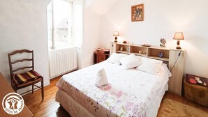 Un dormitorio con una cama grande, dos lámparas de noche, una mesita de madera, una silla y un ventanal con vistas a un edificio.