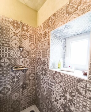 Un baño con baldosas estampadas, ducha, ventana con vistas y un estante pequeño con artículos de aseo.