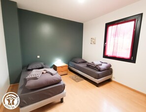 Two single beds with pillows and blankets, a nightstand with a lamp, and a large window with curtains.