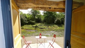 An open door to a patio area with a table and two chairs, a lantern, and a view of a natural landscape.