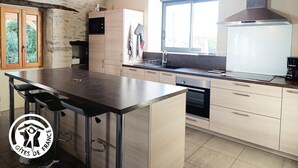 A modern kitchen with a central island, wooden beams, a large window, and a stone wall.