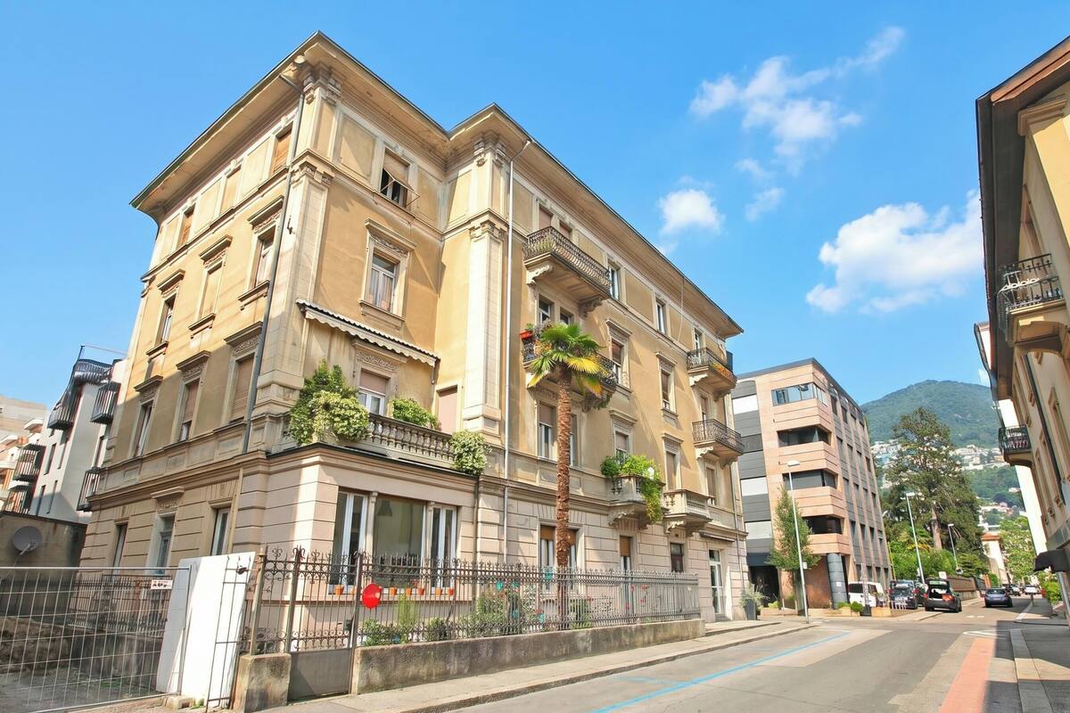 A multi-story building with balconies, a gated entrance, and a clear view of the street and mountains in the background.