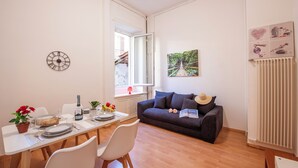 Et spiseområde med et bord dækket til to personer, en sofa og et vindue med udsigt til en bygning.