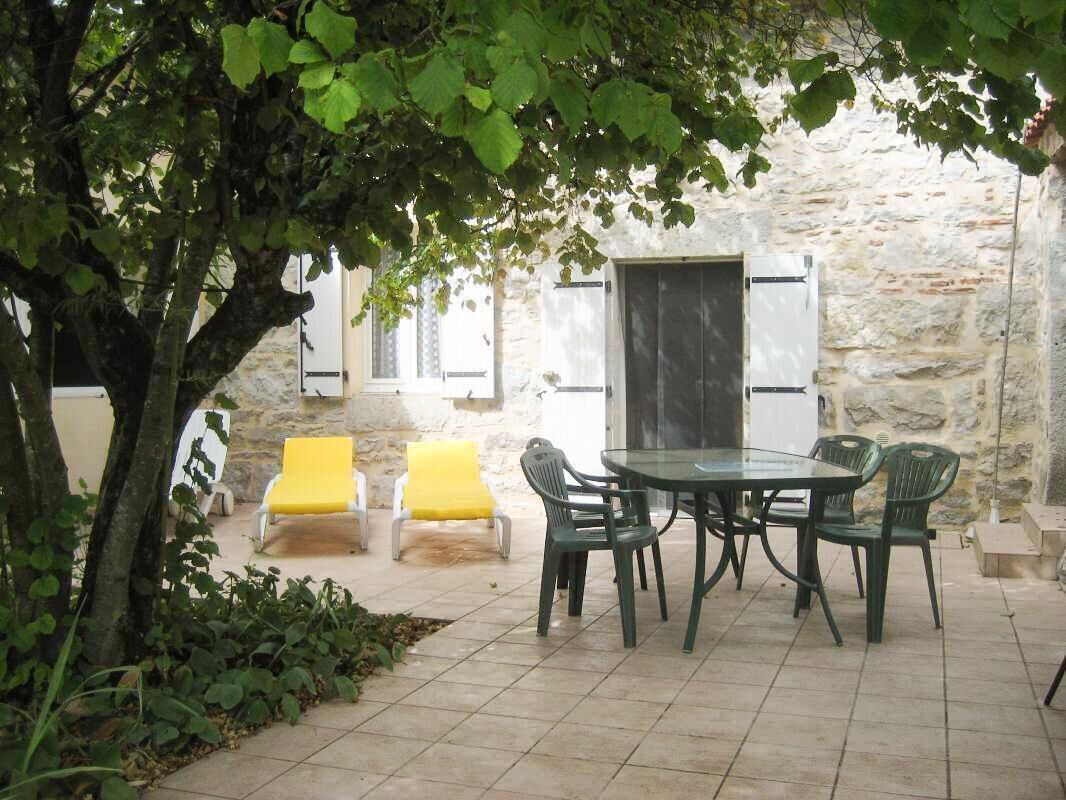 An outdoor patio with a round table and chairs, surrounded by stone walls and a tree providing shade.