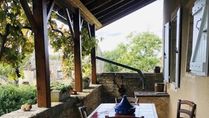 A covered patio with a table and chairs, stone walls, and a view of greenery and buildings.
