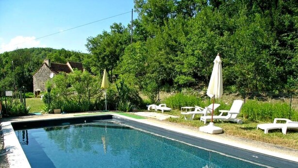 A rectangular swimming pool with clear blue water, surrounded by white lounge chairs and umbrellas, set against a backdrop of lush greenery and a stone building. - 46g12089 - Maxou - Gîte (MAXOU)
