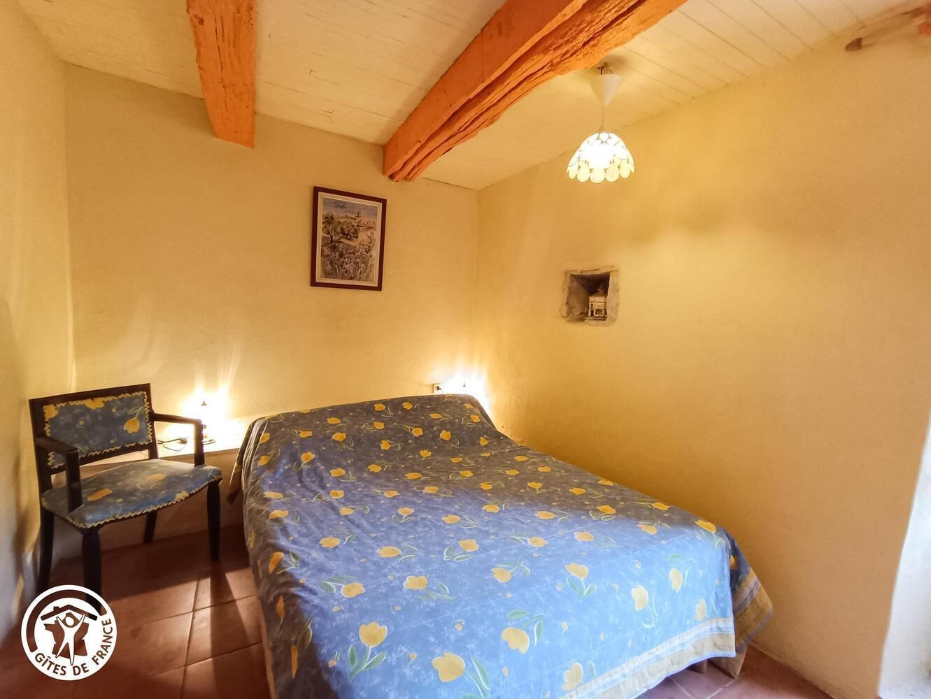 A bedroom with a bed, a chair, a wall-mounted light, and a framed picture.