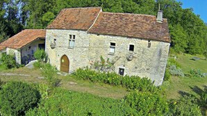 A stone house with a wooden door, a small attached structure, and a garden with yellow flowers. - 46g12089 - Maxou - Gîte (MAXOU)
