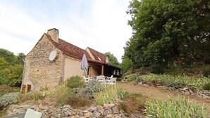 A stone house with a tiled roof, a covered patio with chairs, and a garden with various plants and a stone wall. - 46g12089 - Maxou - Gîte (MAXOU)