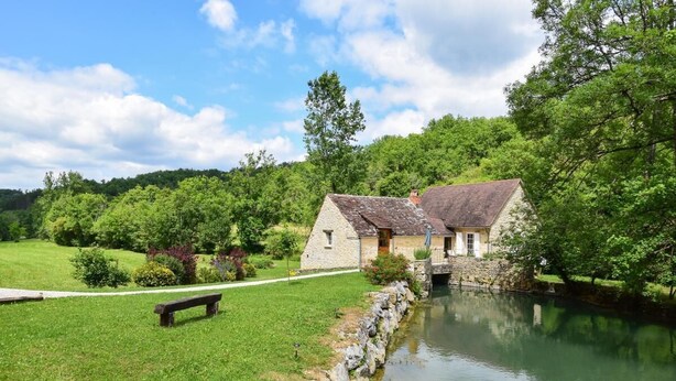 Ein Steinhaus mit einer kleinen Brücke über einen Kanal, umgeben von üppigem Grün und einer Rasenfläche mit einer Bank.