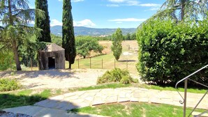 A stone pathway leading to a rustic stone structure, surrounded by lush greenery and tall trees, with a clear view of distant hills. - Borgo Melograno - SOL Apartment (Toscana)