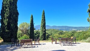 A gravel area with wooden benches and tables, surrounded by tall cypress trees, overlooking a scenic landscape with hills and a clear blue sky. - Borgo Melograno - SOL Apartment (Toscana)