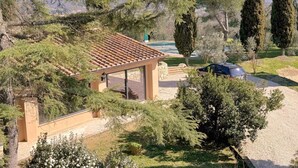 Un espacio de estar al aire libre cubierto con una pérgola, rodeado de árboles y vistas a una piscina y montañas.