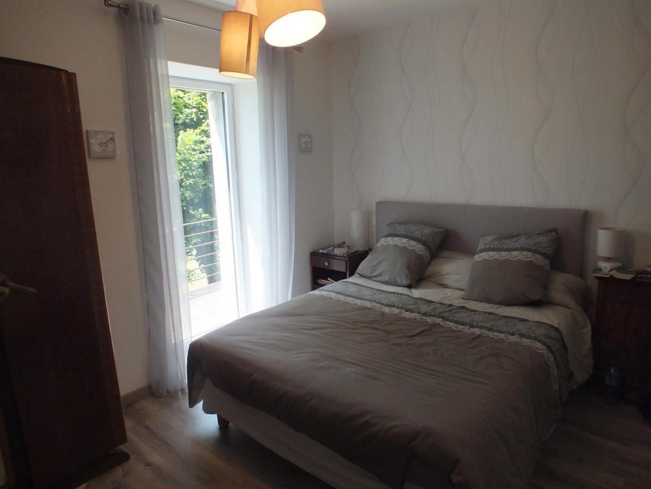 A bedroom with a bed, nightstands, a lamp, and a view of greenery outside.