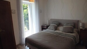 Ein Schlafzimmer mit Bett, Nachttischen, einer Lampe und Blick auf Grünfläche.