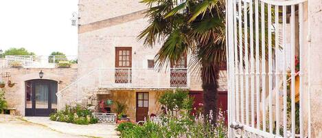 Un edificio de piedra de dos plantas con balcón, una palmera y un portón blanco.