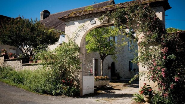 A stone building with a rustic entrance, surrounded by lush greenery and flowering plants. - Gîtes de France® - Chez Mamie (LACAVE)