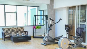 A spacious living room with a large window, a sofa, a coffee table, and exercise equipment including an elliptical and a stationary bike. - Sunrise A Boutique Hotel (Hyderabad)