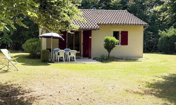 Un petit bâtiment avec un toit en tuiles, une porte et un petit jardin agrémenté d’une pelouse et de quelques chaises.