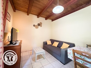 A room with a wooden ceiling, a grey sofa with yellow cushions, a small white table, a TV on a wooden stand, and a logo on the wall. - Gîtes de France® - La Pinède (SOUSCEYRAC EN QUERCY)