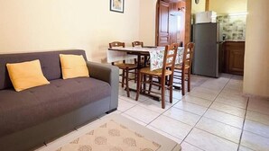 A living room with a sofa and chairs, a dining table with chairs, a kitchen area with a refrigerator, and a tiled floor.