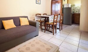 Un soggiorno con divano e poltrone, un tavolo da pranzo con sedie, una zona cucina con frigorifero e un pavimento piastrellato.