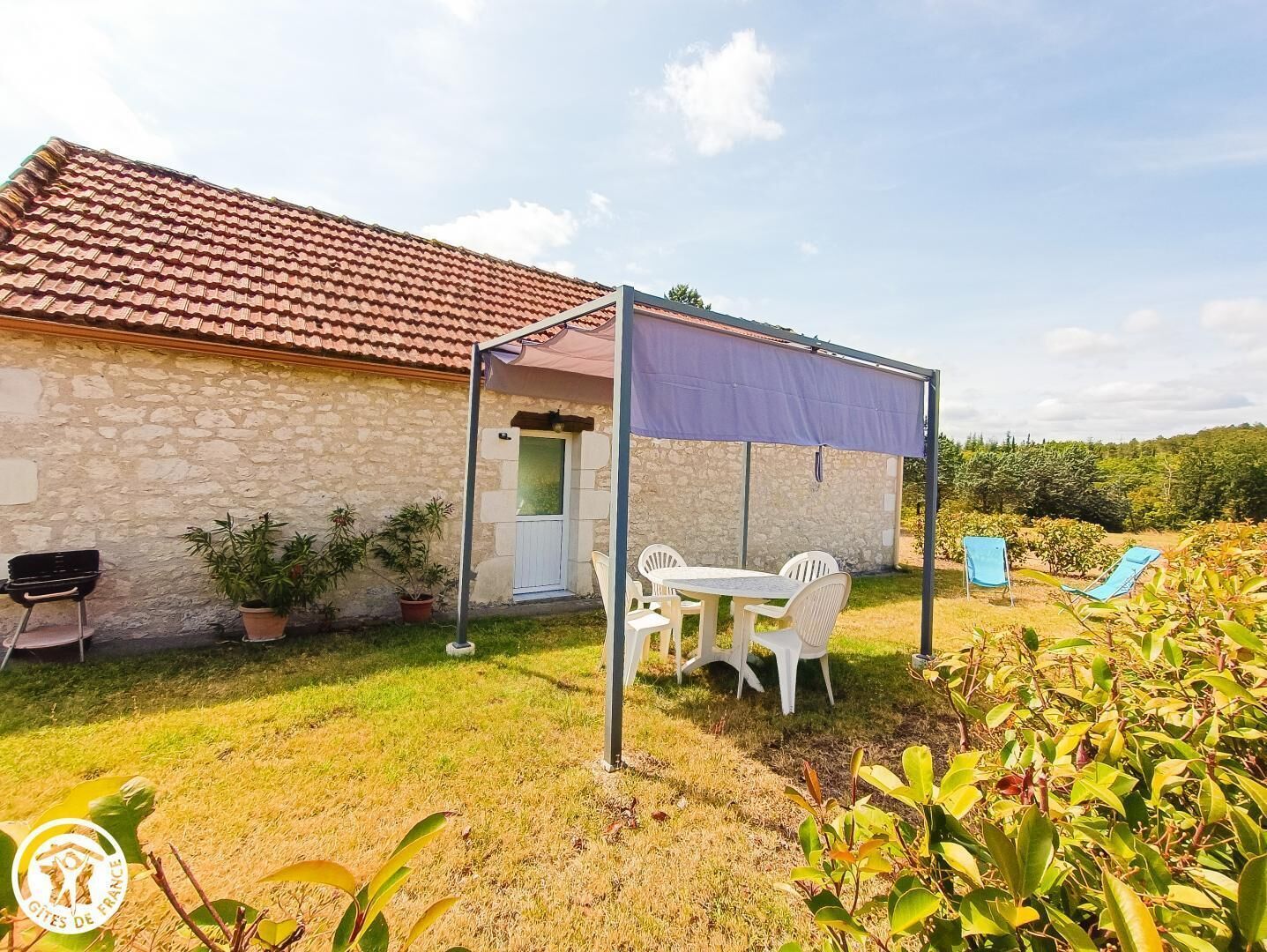 Une maison en pierre avec un toit en tuiles, une terrasse couverte avec des chaises blanches et un petit jardin.