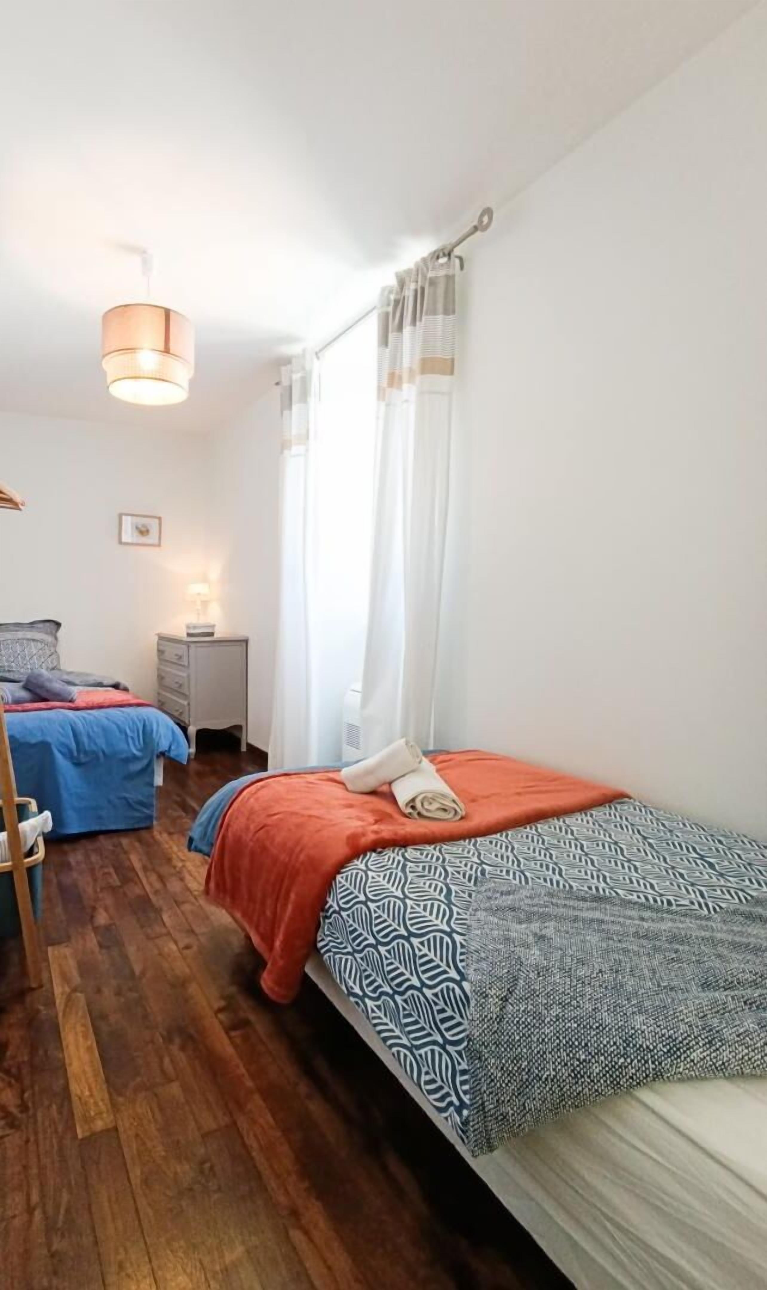 A bedroom with two beds, a wooden floor, a bedside table with a lamp, and a window with curtains.