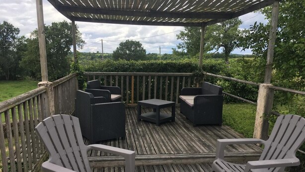 Eine Holzterrasse mit Gartenmöbeln, darunter Stühle und ein Tisch, umgeben von einem Hollerzaun.