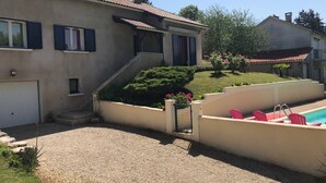 Une maison avec une allée en gravier, un petit jardin agrémenté de fleurs roses et une piscine avec des chaises roses.