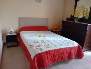 Een net opgemaakt bed met een quilt met bloemenprint, een rode sprei en een nachtkastje met een lamp.