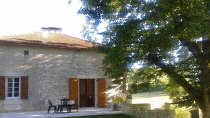 Ein Steinhaus mit Ziegeldach, eine kleine Außenbereich-Sitzgelegenheit mit Stühlen und Tisch sowie ein Baum im Vordergrund.